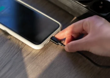 Mão conectando carregador em smartphone sobre mesa de madeira, representando um hábito comum de recarga que pode afetar a vida útil da bateria.