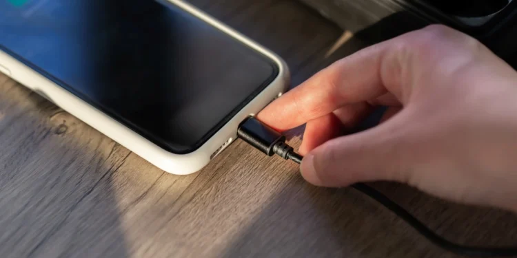 Mão conectando carregador em smartphone sobre mesa de madeira, representando um hábito comum de recarga que pode afetar a vida útil da bateria.