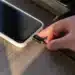 Mão conectando carregador em smartphone sobre mesa de madeira, representando um hábito comum de recarga que pode afetar a vida útil da bateria.