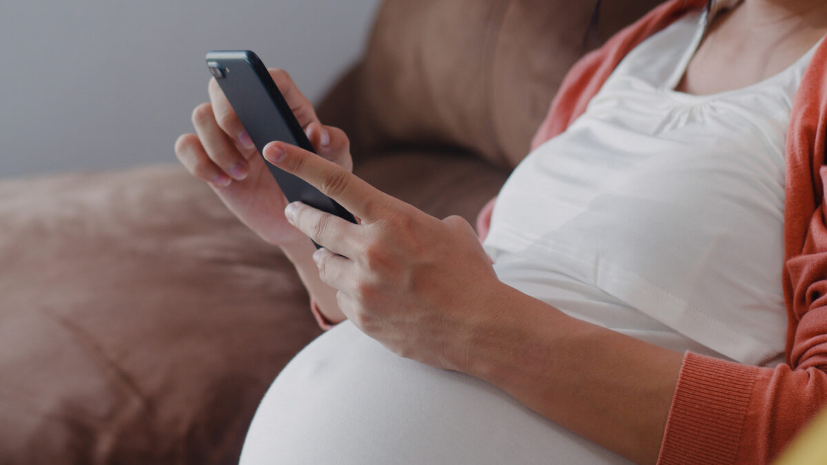 Mulher grávida sentada no sofá usa um celular com as duas mãos.