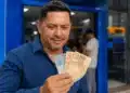 Homem sorrindo ao segurar notas de R$ 100 e R$ 50 em frente a uma agência da Caixa, representando a solicitação do saque a partir desta sexta-feira (29).