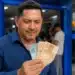Homem sorrindo ao segurar notas de R$ 100 e R$ 50 em frente a uma agência da Caixa, representando a solicitação do saque a partir desta sexta-feira (29).