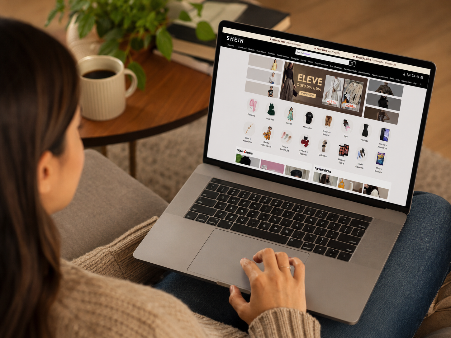 Jovem de suéter bege usa MacBook para acessar loja virtual SHEIN Brasil, exibindo categorias de moda feminina