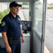 Vigilante patrimonial uniformizado em guarita com monitor de CFTV, cancela e livro de registro ao fundo.