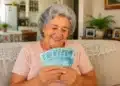 Senhora sorrindo ao conferir cédulas de reais em sua sala, simbolizando pagamento da segunda parcela do 13º salário.