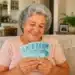 Senhora sorrindo ao conferir cédulas de reais em sua sala, simbolizando pagamento da segunda parcela do 13º salário.