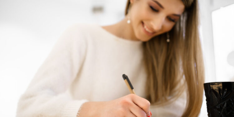 Jovem mulher de blusa branca sorridente escrevendo em caderno aberto sobre mesa clara