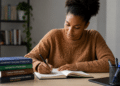 Mulher negra de suéter marrom escreve em caderno com pilha de livros para concurso sobre mesa.