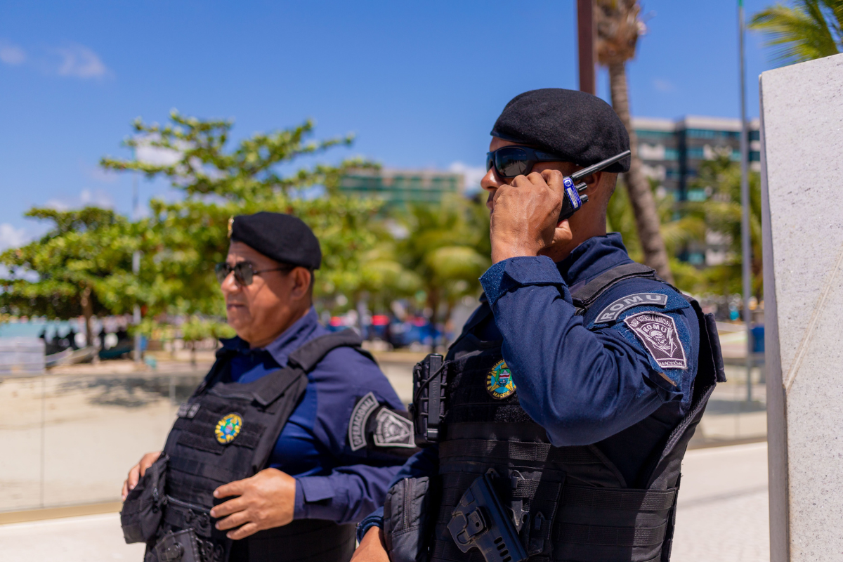 Dois guardas municipais da ROMU Maceió em patrulha, vestindo uniforme azul-marinho com colete tático, boina preta e óculos escuros