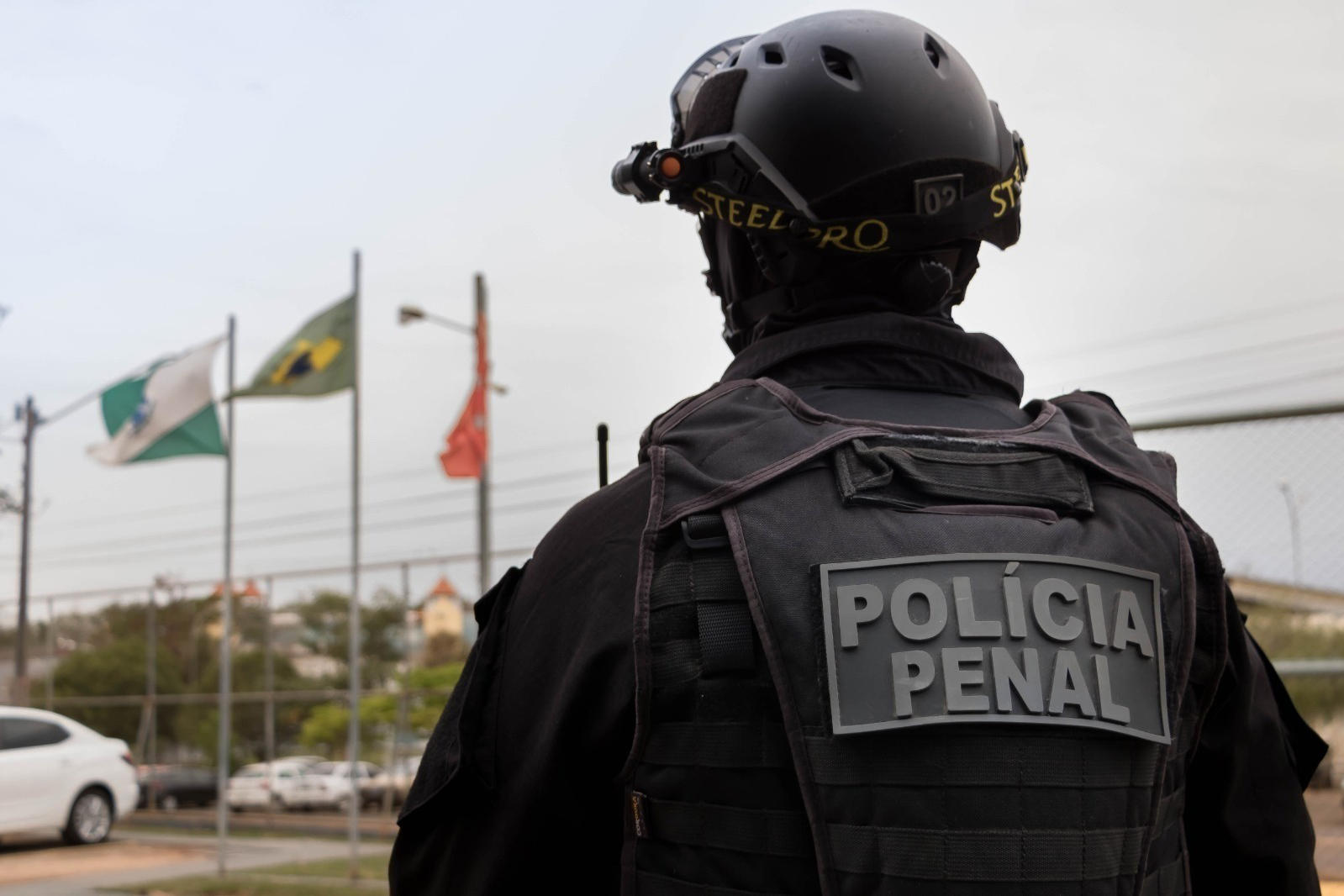 Policial Penal de costas com uniforme tático preto, capacete e óculos de visão noturna, frente a bandeiras do Acre e Brasil