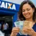 Mulher sorrindo em frente à agência da Caixa Econômica Federal segurando notas de real, representando os R$ 6.220 disponíveis até o fim de maio para CPFs com finais de 0 a 9.