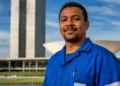 Trabalhador brasileiro com uniforme azul em frente a prédio público, representando a PEC do fim da escala 6x1, que pode ser votada na próxima semana.