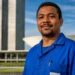 Trabalhador brasileiro com uniforme azul em frente a prédio público, representando a PEC do fim da escala 6x1, que pode ser votada na próxima semana.