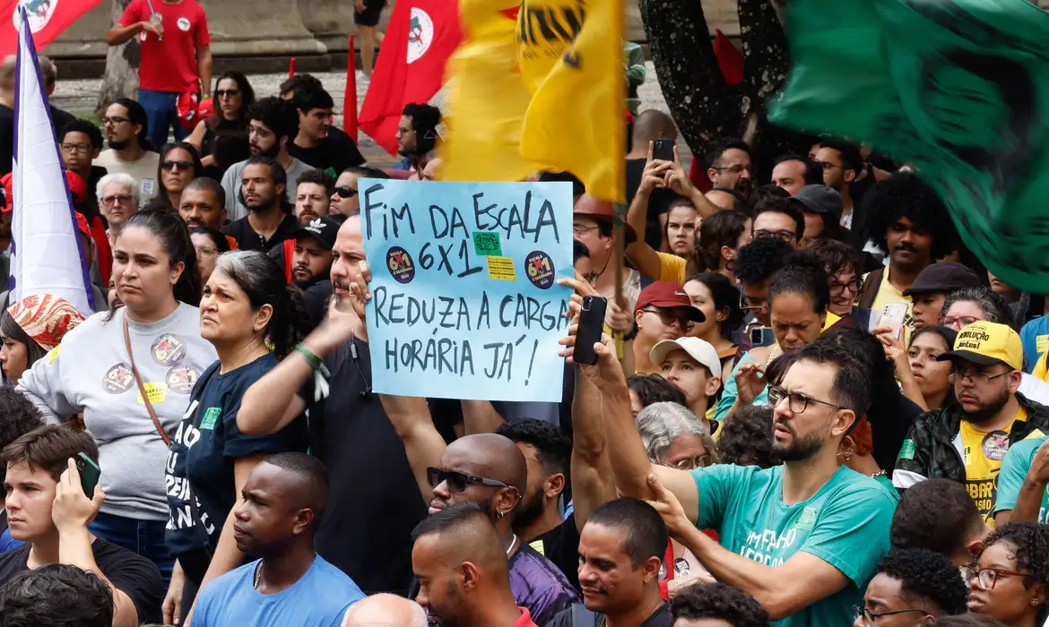 Multidão concentrada em ato público segura cartaz azul com a inscrição fim da escala 6x1 em meio a bandeiras vermelhas e amarelas de movimentos sociais