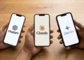 Três smartphones exibindo logos de ChatGPT, Claude e Gemini segurados por mãos sobre mesa de madeira.