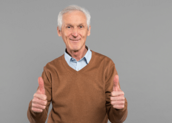 Senhor idoso sorridente faz sinal positivo com as mãos em fundo cinza.