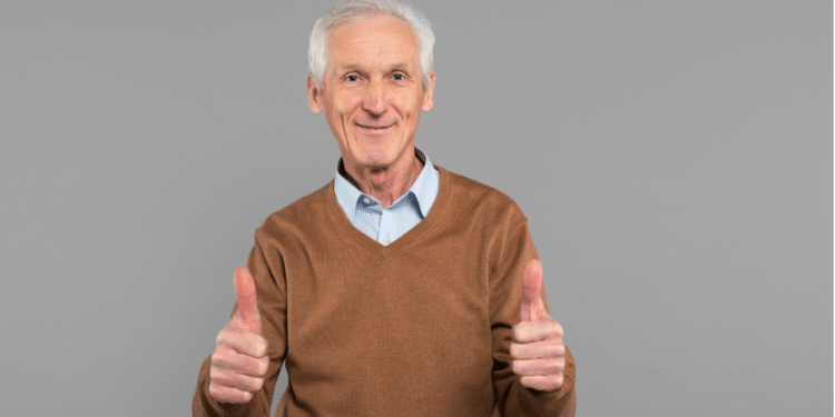 Senhor idoso sorridente faz sinal positivo com as mãos em fundo cinza.