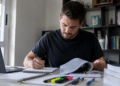 Homem jovem escreve em caderno, consulta livro aberto, com laptop e marca-textos coloridos na mesa.
