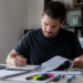 Homem jovem escreve em caderno, consulta livro aberto, com laptop e marca-textos coloridos na mesa.