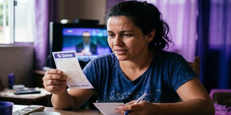 Mulher de camiseta azul segura volante da Quina com caneta na mão, em sala com TV ligada ao fundo