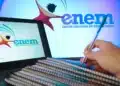 Notebook exibindo logo do Enem, cadernos empilhados e mão segurando caneta em fundo azul com marca do exame