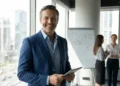 Foto de um profissional de Gestão Comercial sorrindo em escritório, com colegas ao fundo e gráfico de resultados ao lado.