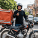 Entregador de aplicativo faz sinal positivo com mochila térmica na moto.