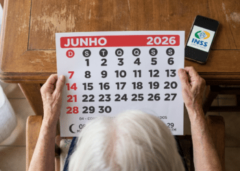 Pessoa idosa com cabelos brancos segura calendário de junho 2026 sobre mesa de madeira, com celular exibindo logo do INSS.