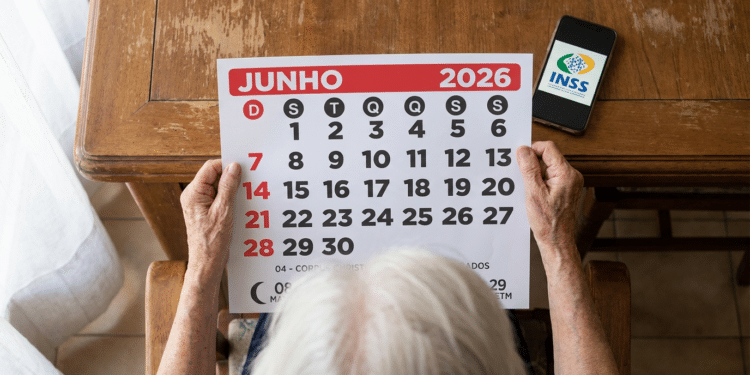 Pessoa idosa com cabelos brancos segura calendário de junho 2026 sobre mesa de madeira, com celular exibindo logo do INSS.