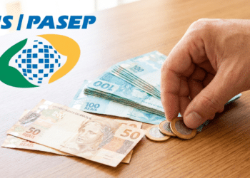 Mão pegando moedas ao lado de notas de reais e logotipo PIS/PASEP sobre mesa de madeira.