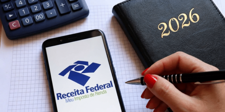 Celular com logo da Receita Federal, calculadora e agenda preta de 2026 ao lado de mão segurando caneta.