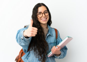 Jovem estudante sorridente com óculos faz sinal de positivo em fundo claro.