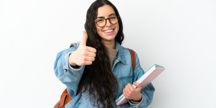 Jovem estudante sorridente com óculos faz sinal de positivo em fundo claro.