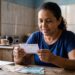 Mulher de camiseta azul segura bilhete da Loteria Federal com notas de reais sobre mesa de madeira