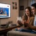 Casal sentado no sofá observa bilhete da Loteria Federal enquanto assiste ao sorteio da Caixa na TV