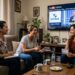 Três mulheres em sala assistem ao sorteio LOTECE 24/06 na TV, sorrindo e conferindo o bilhete.