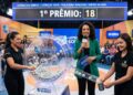Apresentadora anuncia prêmio 18 com assistentes operando o globo da Loteria dos Sonhos em estúdio colorido
