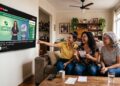 Três mulheres sorridentes acompanham o sorteio da Loteria dos Sonhos na TV e conferem bilhetes em sala iluminada