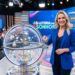 Mulher loira sorrindo revela bola sorteada em estúdio da Loteria dos Sonhos com plateia ao fundo