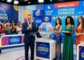 Apresentador anuncia sorteio da Loteria dos Sonhos ao vivo com assistentes em vestidos coloridos e painel digital