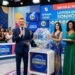 Apresentador anuncia sorteio da Loteria dos Sonhos ao vivo com assistentes em vestidos coloridos e painel digital