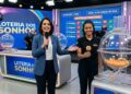 Apresentadora e assistente mostram bola número 7 durante sorteio da Loteria dos Sonhos, painel de resultados ao fundo