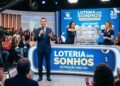 Apresentador anuncia sorteio da Loteria dos Sonhos com duas assistentes e público ao fundo em estúdio iluminado