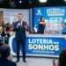 Apresentador anuncia sorteio da Loteria dos Sonhos com duas assistentes e público ao fundo em estúdio iluminado