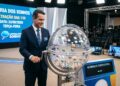 Homem de terno azul operando globo da sorte em estúdio de loteria, painel mostra data e horário