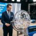 Homem de terno azul operando globo da sorte em estúdio de loteria, painel mostra data e horário