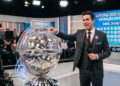 Apresentador de terno ao lado do globo sorteador da Loteria dos Sonhos, com números exibidos em painel e data 26/06/2026