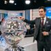 Apresentador de terno ao lado do globo sorteador da Loteria dos Sonhos, com números exibidos em painel e data 26/06/2026