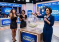 Três apresentadoras apresentam sorteio da Loteria dos Sonhos com bolas numeradas e público ao fundo