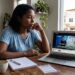 Mulher assiste ao sorteio da Loteria dos Sonhos no notebook, anotando resultados na mesa de casa